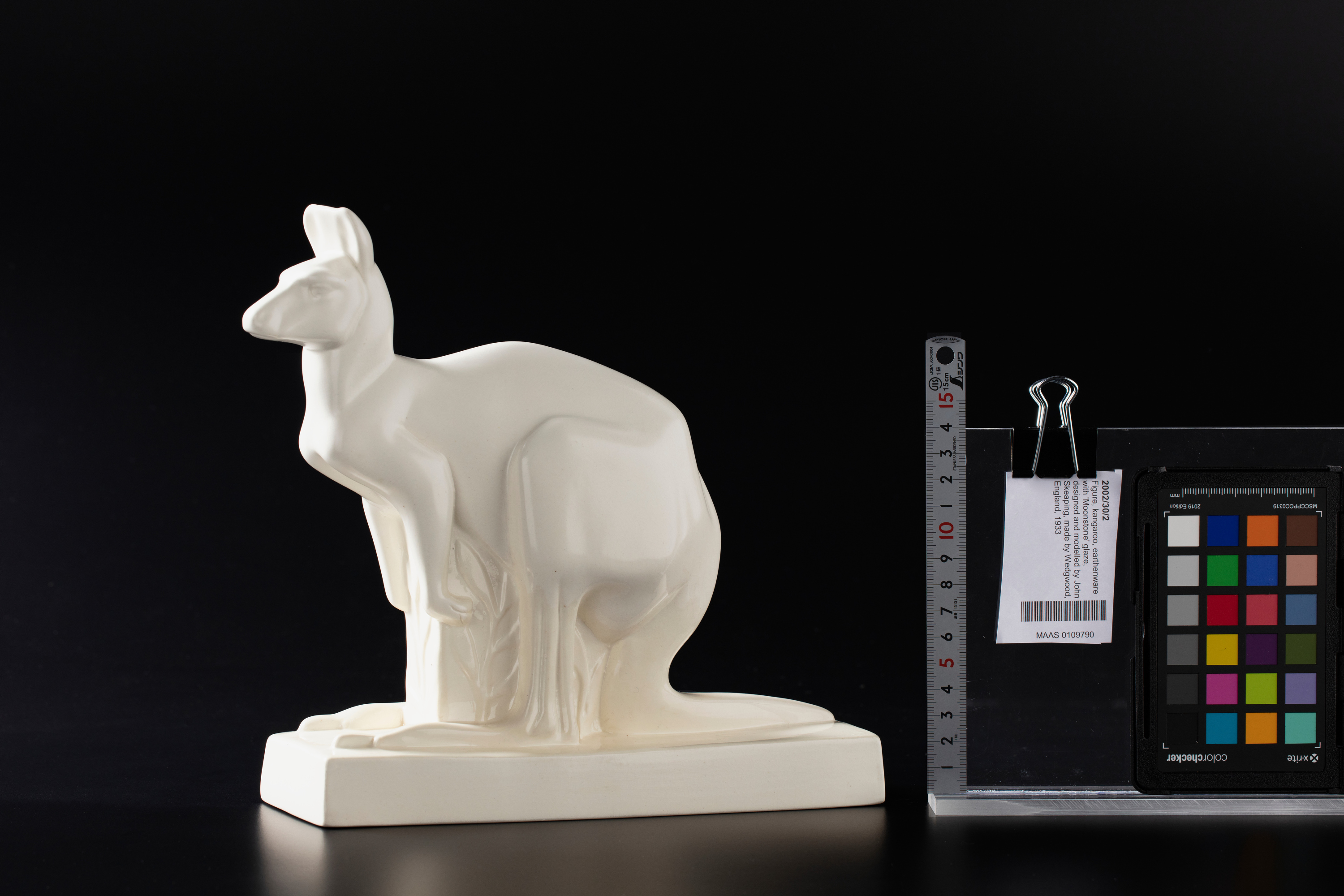 White earthenware kangaroo on a black studio background. To the right of the kangaroo is a upright metal ruler (for scale), paper object tag, and colour reference chart. The kangaroo is about 20cm tall.